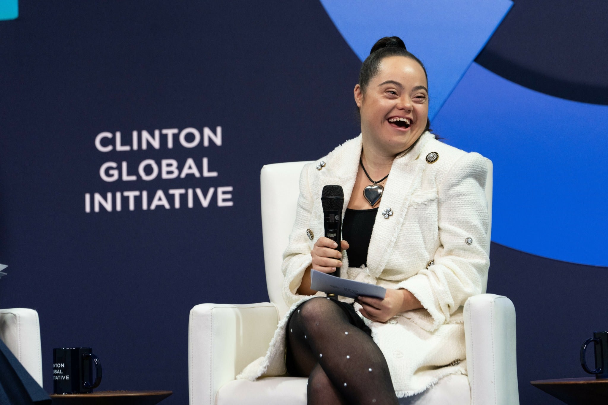Madison smiling brightly while speaking onstage at the Clinton Global Initiative event, holding a microphone.