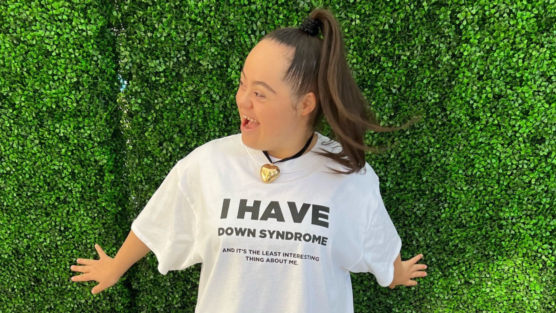 Madison wearing a white shirt that says “I Have Down Syndrome, and it's the least interesting thing about me” standing in front of a green backdrop and smiling.